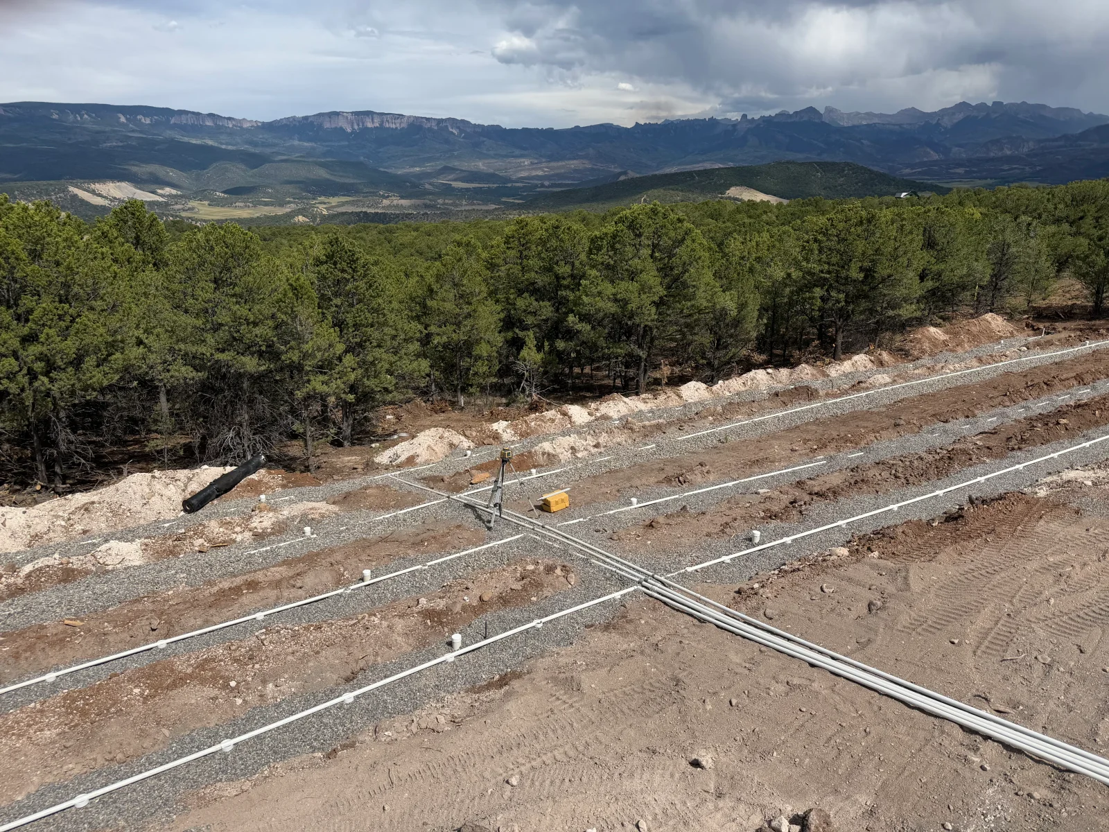 Graded building site with utility trenches, cleared timber, and mountain forest backdrop